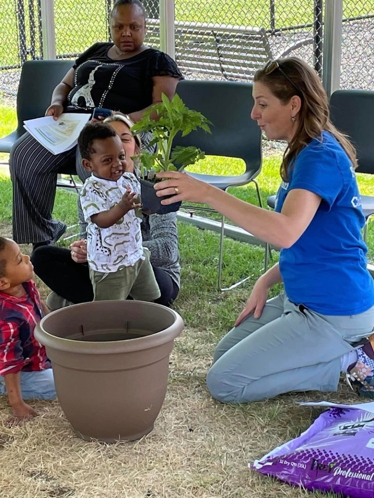 A woman in a T-shirt offers a plant to a toddler next to a gardening pot.