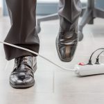 Feet of someone who is walking in men’s dress shoes, in an office setting, about to trip over the cord from a power strip.