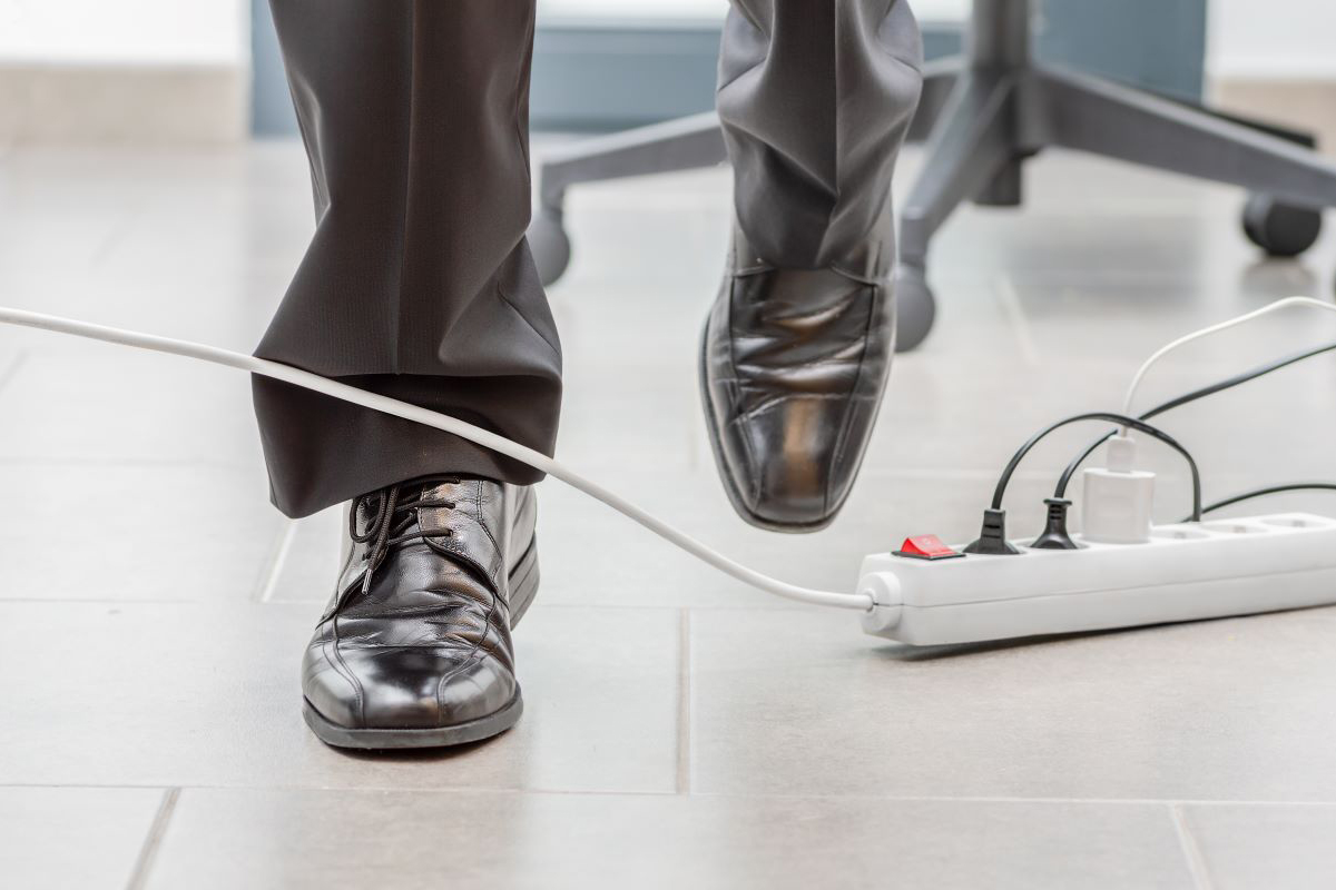 Feet of someone who is walking in men’s dress shoes, in an office setting, about to trip over the cord from a power strip.