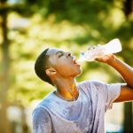 Young man sweating, drinking water from a bottle outside on a sunny day.