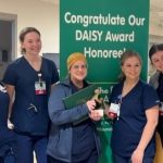 Five people stand in a row in front of a sign that says "Congratulate Our Daisy Award Honoree." The woman in the center holds a plaque.