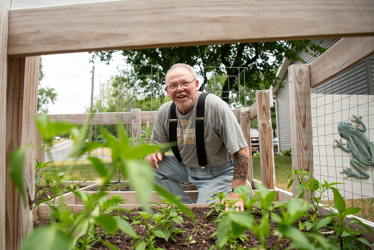 John Kline, wearing suspenders and glasses, kneels on the ground as he looks over his garden. Plants are visible in wooden planter beds. In the background are trees and a building on the right. In the far-right side of the photo, a decorative frog hangs on the garden door.