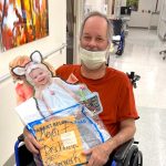 Man wearing a medical mask, seated in a wheel chair, holds an enlarged cardboard cutout of a photo of a little girl in an Easter costume and his hospital discharge paperwork.
