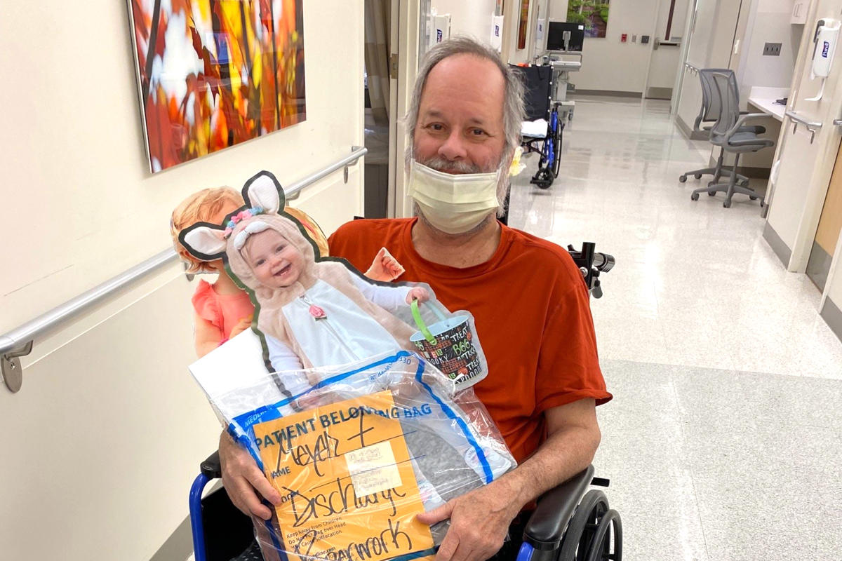 Man wearing a medical mask, seated in a wheel chair, holds an enlarged cardboard cutout of a photo of a little girl in an Easter costume and his hospital discharge paperwork.