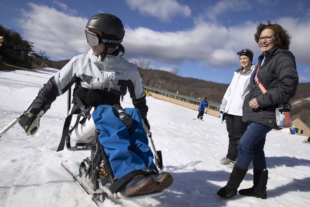 Two women and smile and laugh next to a man on an adaptive ski. They stand on a fine bed of snow.