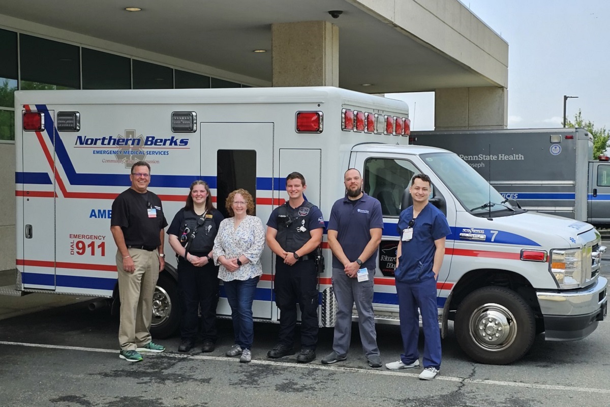 Six adults stand in front of an ambulance parked in front of a hospital. A second ambulance is visible behind it to the right. The man standing on the left and the two men on the right have name badges clipped to their clothing. The woman second from left and the man fourth from left are wearing vests outfitted with a radio/intercom system. The woman has sunglasses clipped to her vest.