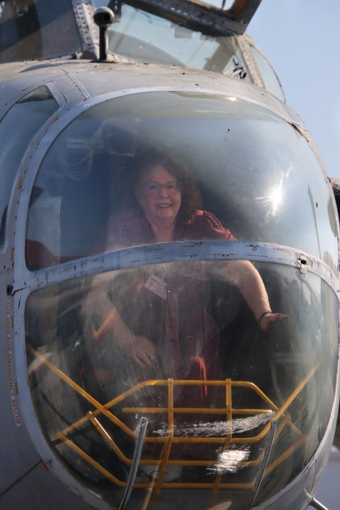 A woman sits in the cockpit of a World War II helicopter. She is wearing glasses and a name badge is clipped to her clothing. Looped wires are hanging from an instrument to the left of her head. A series of metal bars are visible at the bottom of the cockpit. 