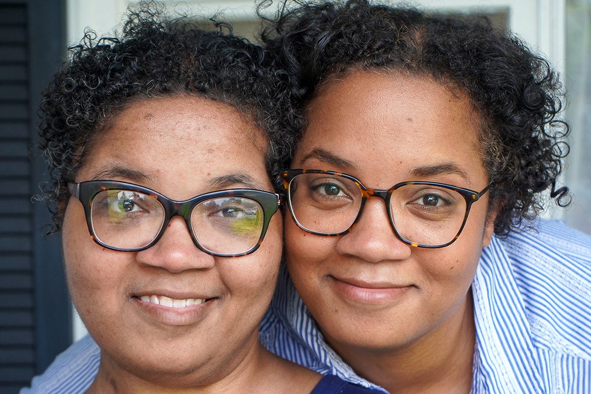 Twin sisters Alana and Alicia Webb smile as they pose in front of a window at their home. Both women have short curly hair and wear glasses and blue shirts.