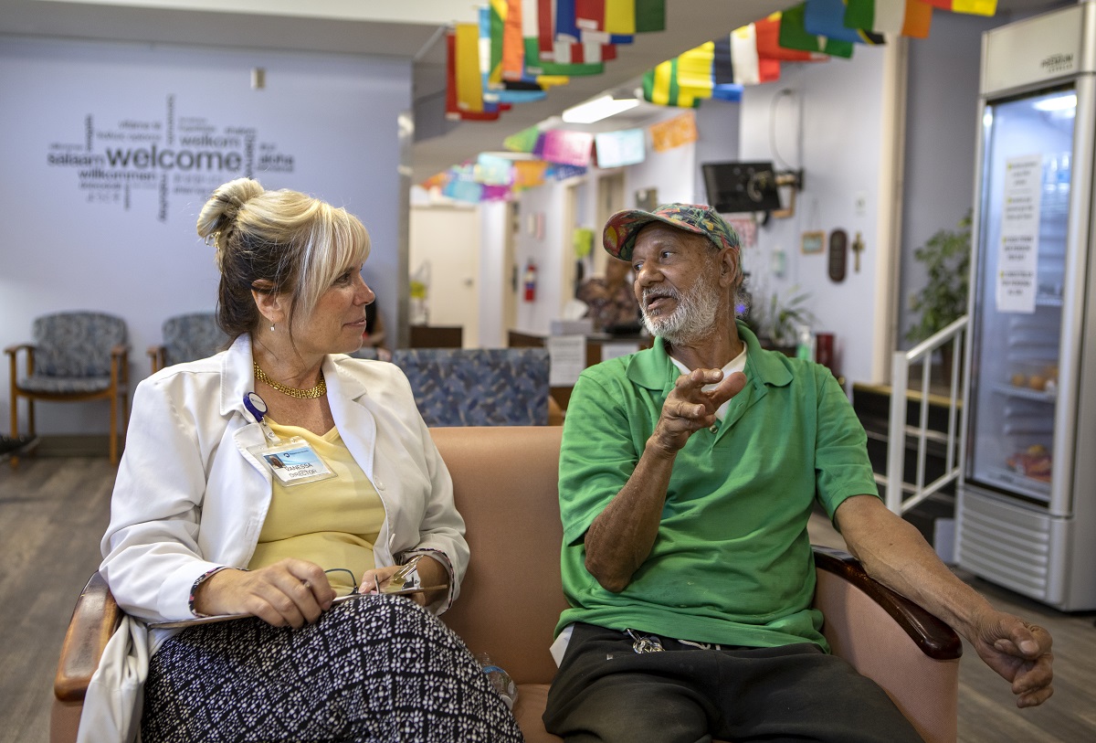 A man and a woman sit side by side on a couch in a waiting room. Overhead, flags from many different countries are attached to the ceiling.