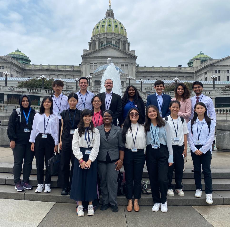 Penn Staters and international students enrolled in Penn State College of Medicine’s Global Health Exchange Program stand in a group outside on the steps of the State Capitol in Harrisburg as part of their three-week immersive training program. A fountain and green-domed building can be seen in the background.
