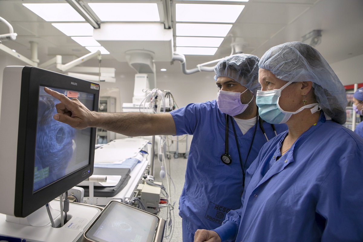 Dr. Vasudev Virparia, left, points at an intravascular ultrasound image on a monitor while Norah Martin, a registered cardiovascular invasive specialist at Penn State Health Lancaster Medical Center, watches. They are both wearing scrubs, face masks and surgical caps. They are standing in a cardiac catheterization lab.