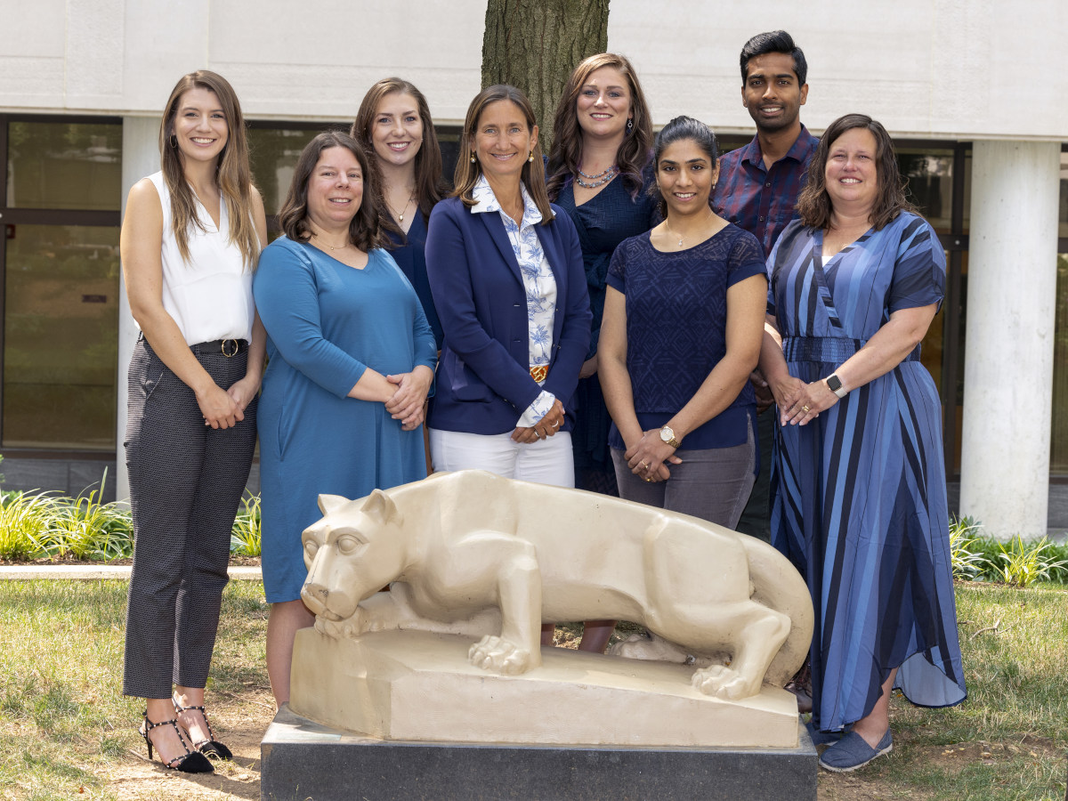 Eight people in casual business attire pose behind a Nittany Lion statue. A tree as well as a building made of concrete and windows is in the background.