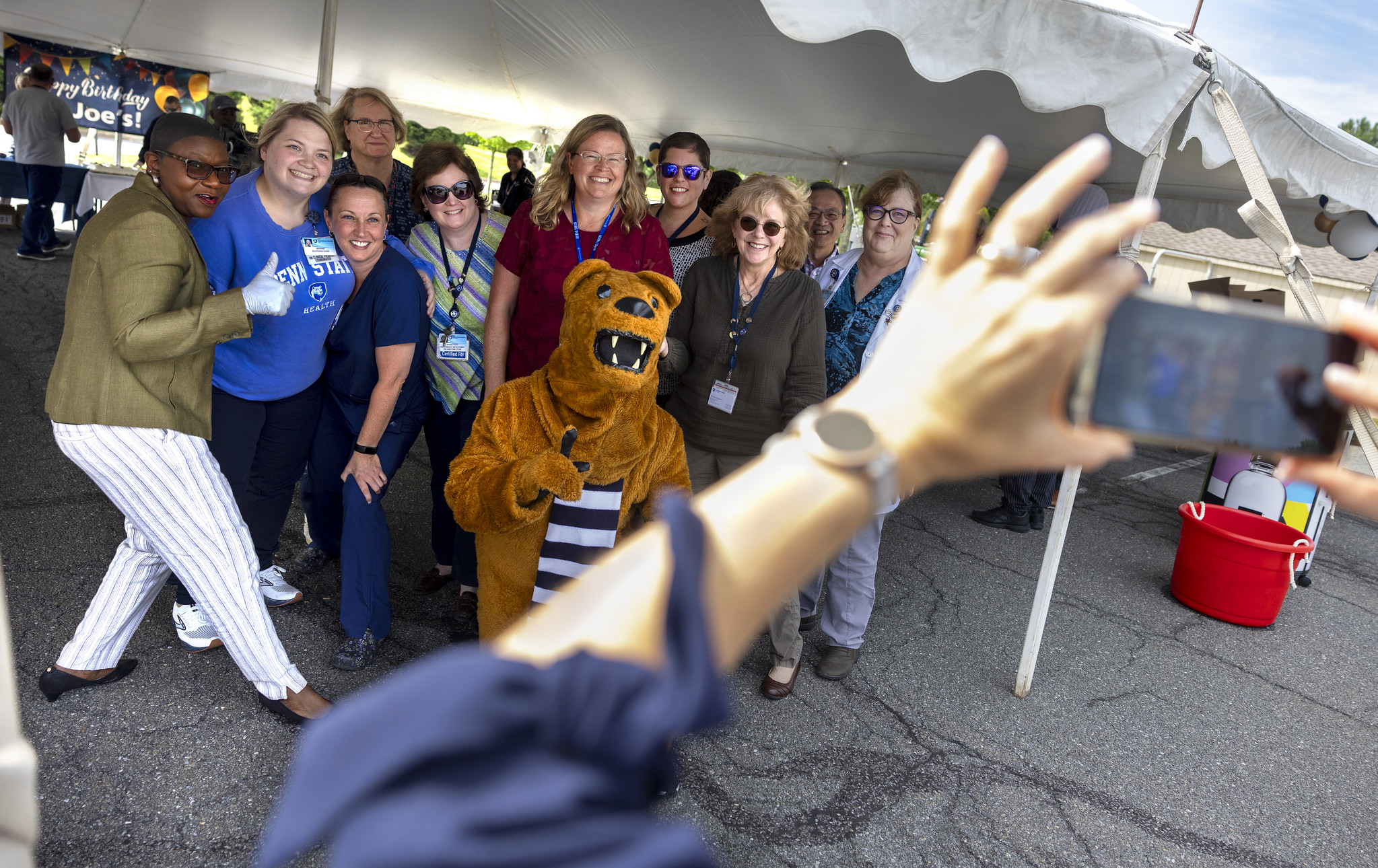 A group of about 10 people pose for a photo with the Penn State Nittany Lion underneath an outdoor event tent.