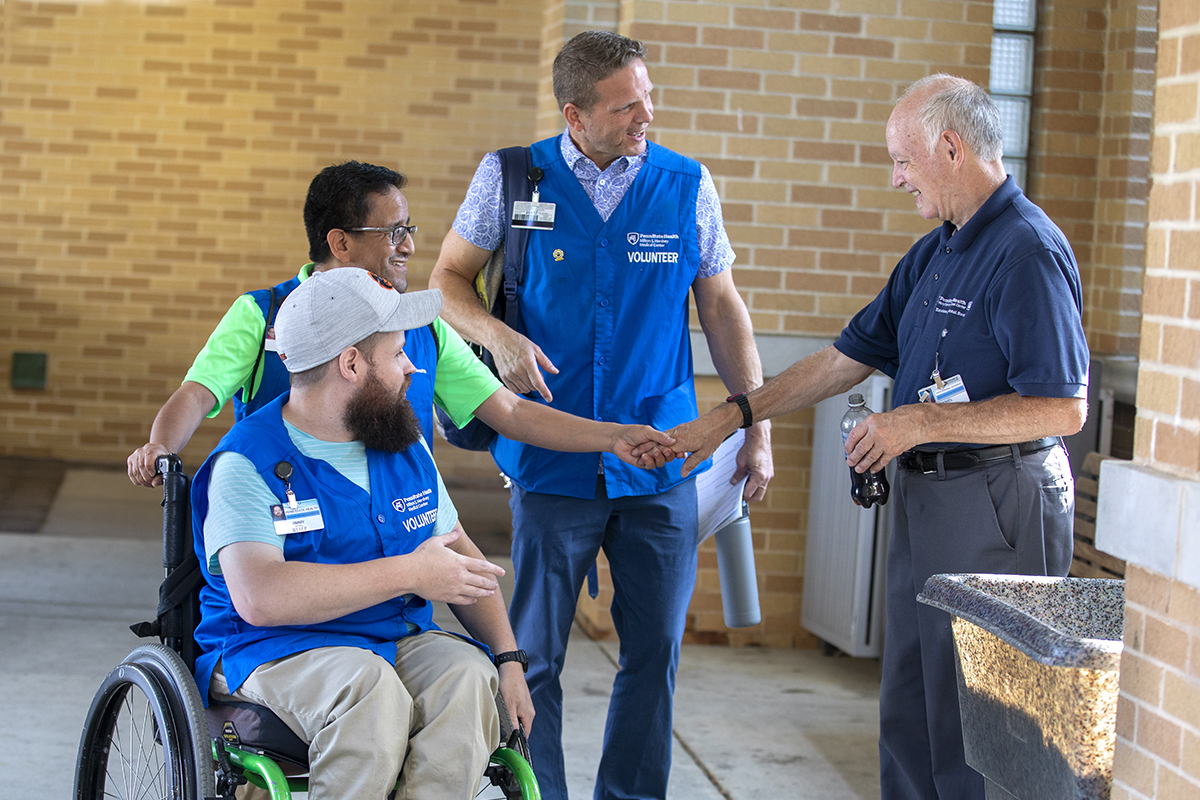 Three men in blue hospital volunteer vests, one in a wheelchair, talk to a male hospital employee in a hospital entryway.