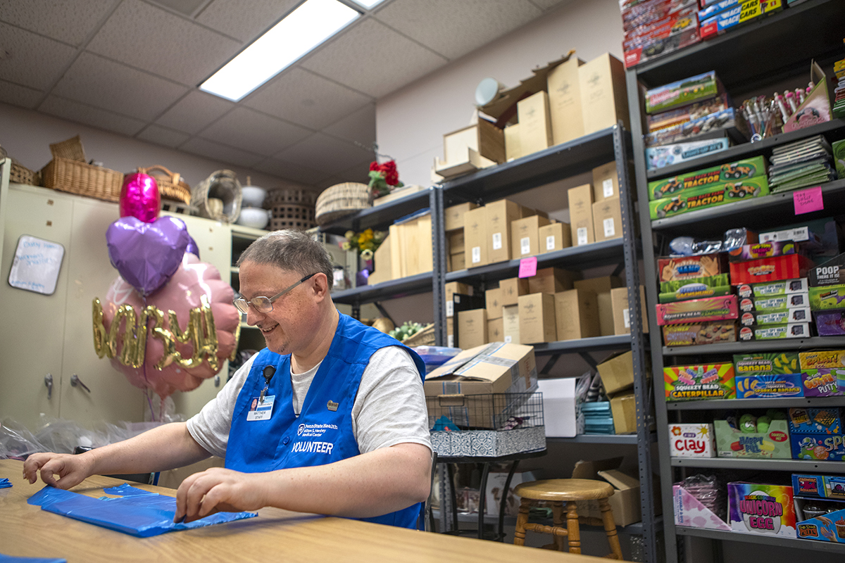 A man wearing a blue hospital volunteer vest is seated at a table organizing bags in a hospital gift shop stock room.