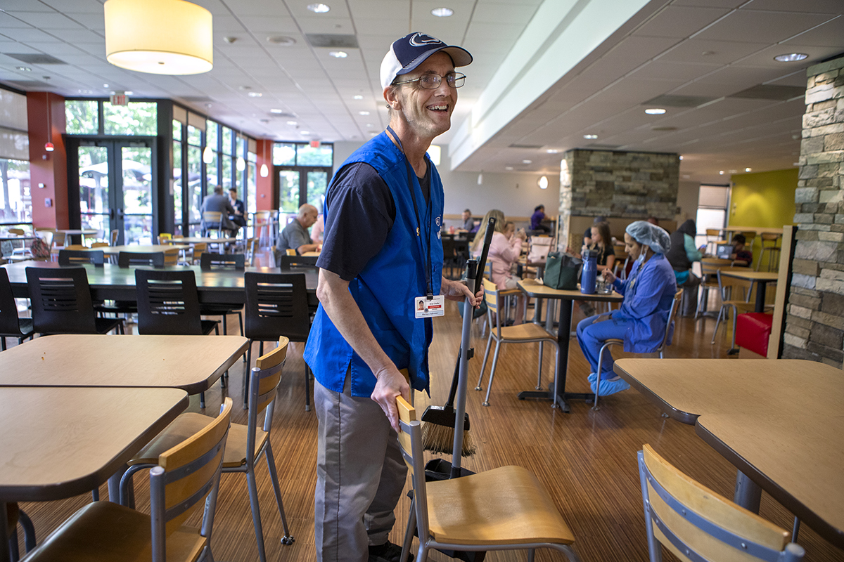 A man wearing a blue hospital volunteer vest sweeps a floor in hospital cafeteria.
