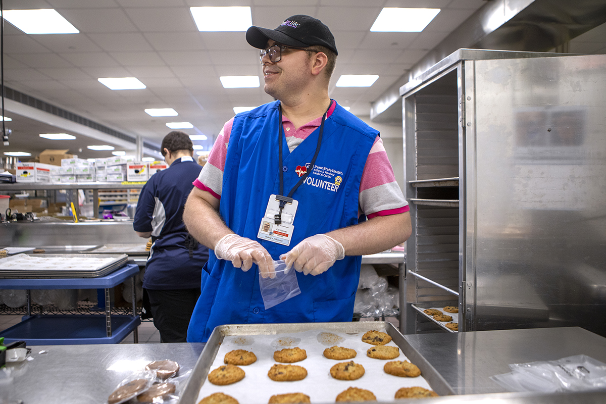 A man wearing a blue hospital volunteer vest sorts cookie in a hospital kitchen.