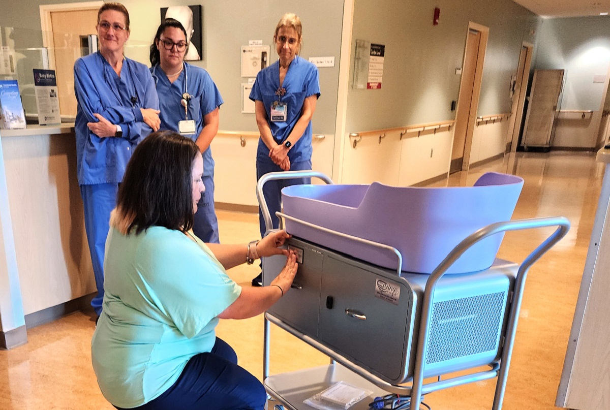 A kneeling woman touches a metal plate on the side of a cart that sits at the entrance of a hospital hallway. A bassinet sits on top of the cart. An electrical cord and a bag of wipes sit on the lower shelf of the cart. Three nurses stand in the background. Posters and railings hang on the hospital walls. In the hallway on the right are two doorways. A tall cart sits in the corner at the end of the hallway.