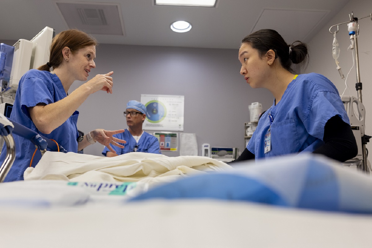 Two women lean across a hospital bed. The woman on the left gestures at the woman on the right. A man in the background looks on.