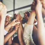 Close-up of diverse people’s arms raised and clasping hands. Professional environment is implied by background and professional clothing sleeves.