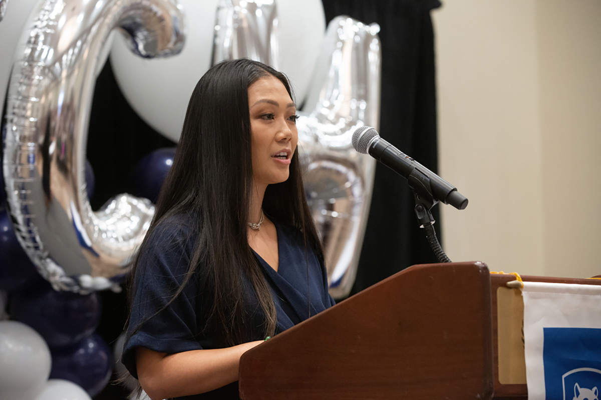 Woman with long hair speaking at a podium with a microphone, letter-shaped balloons in the background.
