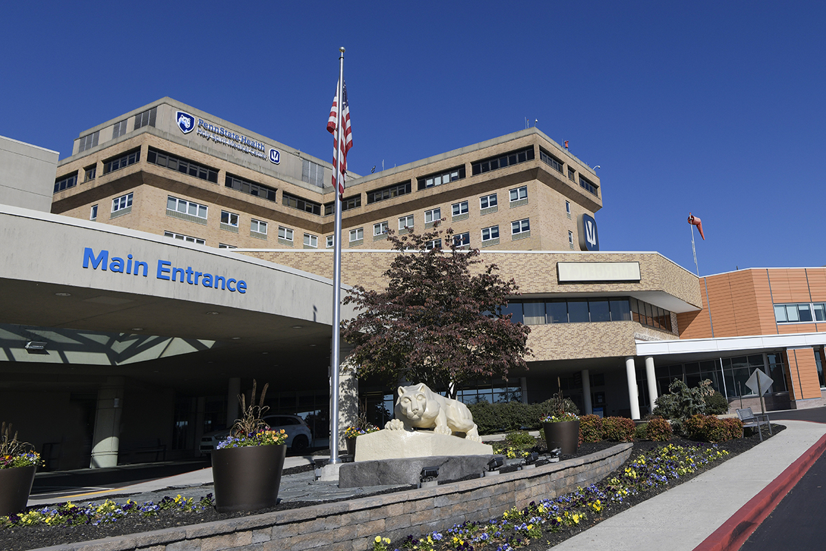 Hospital building: Small wall of pavers lined with flowers, Nittany Lion statue, American flag in foreground; building in background with 