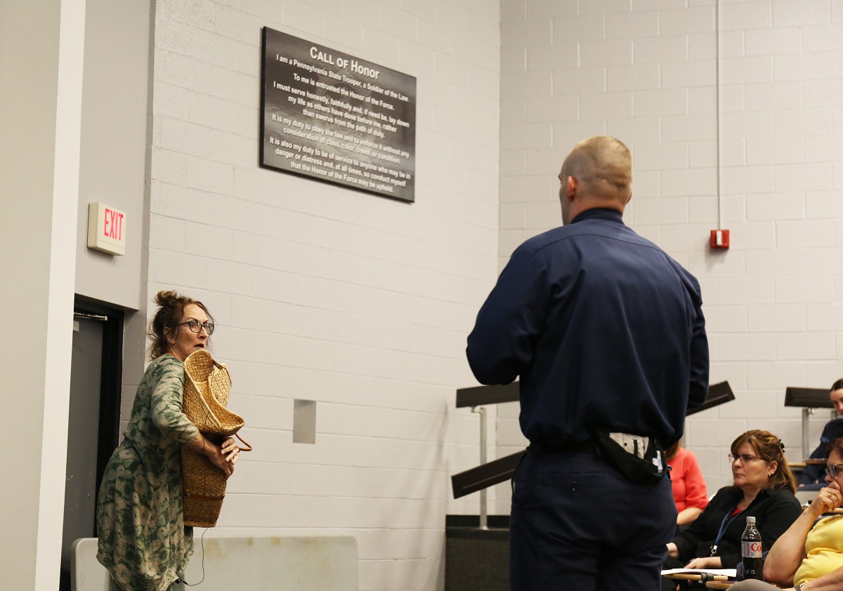 A woman holds a wicker basket to her chest and looks at a police officer with a frightened expression on her face. She is wearing a flowered dress and glasses. The back of a police officer is visible on the right. Four people sit at desks and observe.