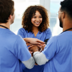 Four health care workers wearing scrubs standing in a circle facing each other, stacking their hands with each other’s, smiling.