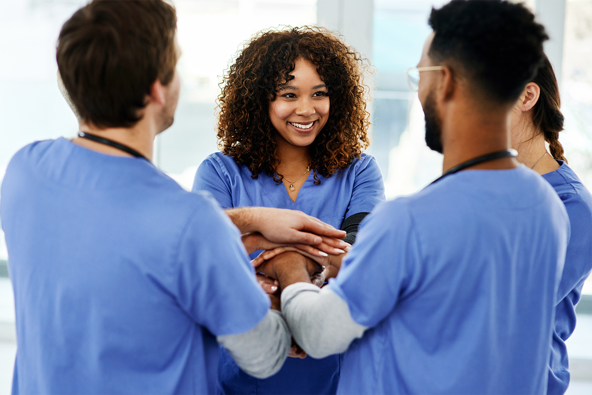 Four health care workers wearing scrubs standing in a circle facing each other, stacking their hands with each other’s, smiling.