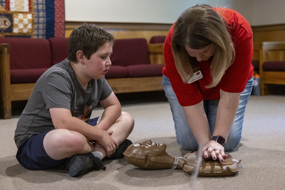 A young boy watches a woman perform chest compressions on a medical doll.