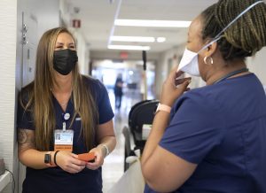 Kristen Hays, left, talks to her team member, Danielle Evans. They are both wearing scrubs and face masks. Hays has long hair and a name badge hanging from her blouse.
