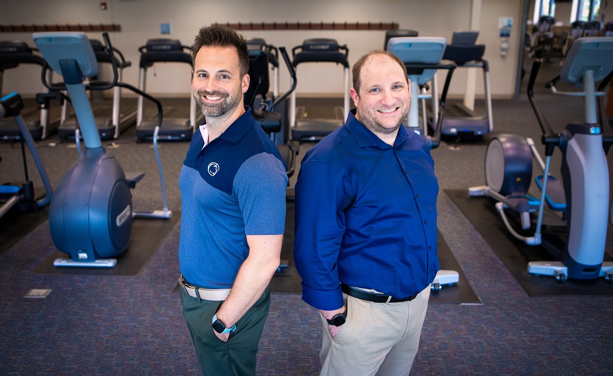 Two men stand in front of exercise equipment.