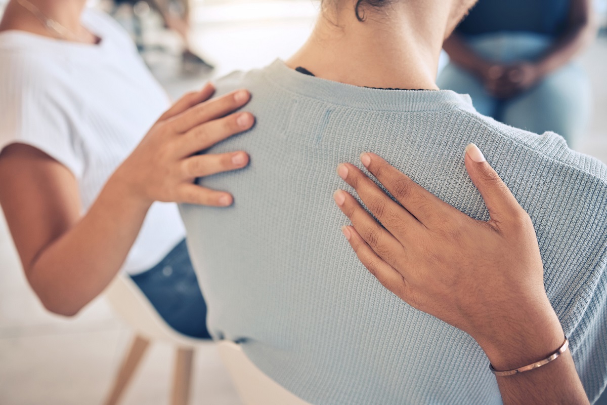 People in a group therapy session put their hands on a woman’s back to show support.