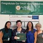 Four ladies, one holding a certificate portfolio, all smiling. On the wall behind them, a bulletin board and a sign that says, “Congratulate Our DAISY Award Honoree!”
