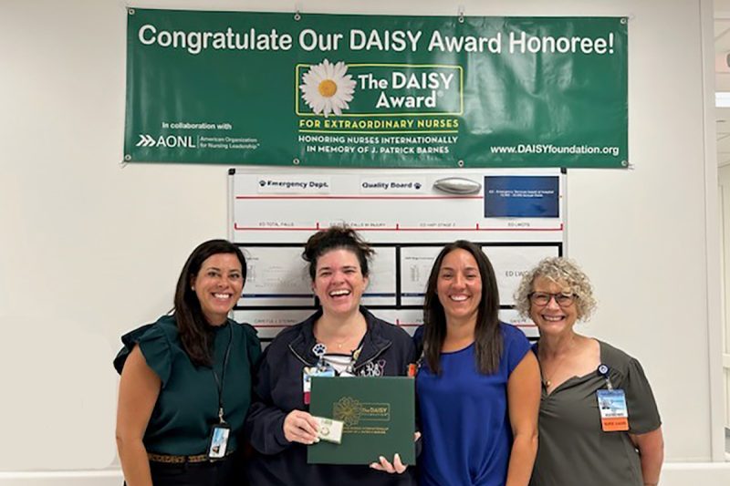 Four ladies, one holding a certificate portfolio, all smiling. On the wall behind them, a bulletin board and a sign that says, “Congratulate Our DAISY Award Honoree!”