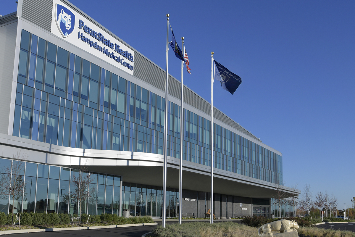 Hospital building with large, reflective windows on most of the exterior, a sign that says, “Penn State Health Hampden Medical Center” with a shield containing the image of a mountain lion – The Penn State Nittany Lion. Three flags in front of the building – American, Pennsylvania, Penn State Health. Nittany Lion statue in the foreground.