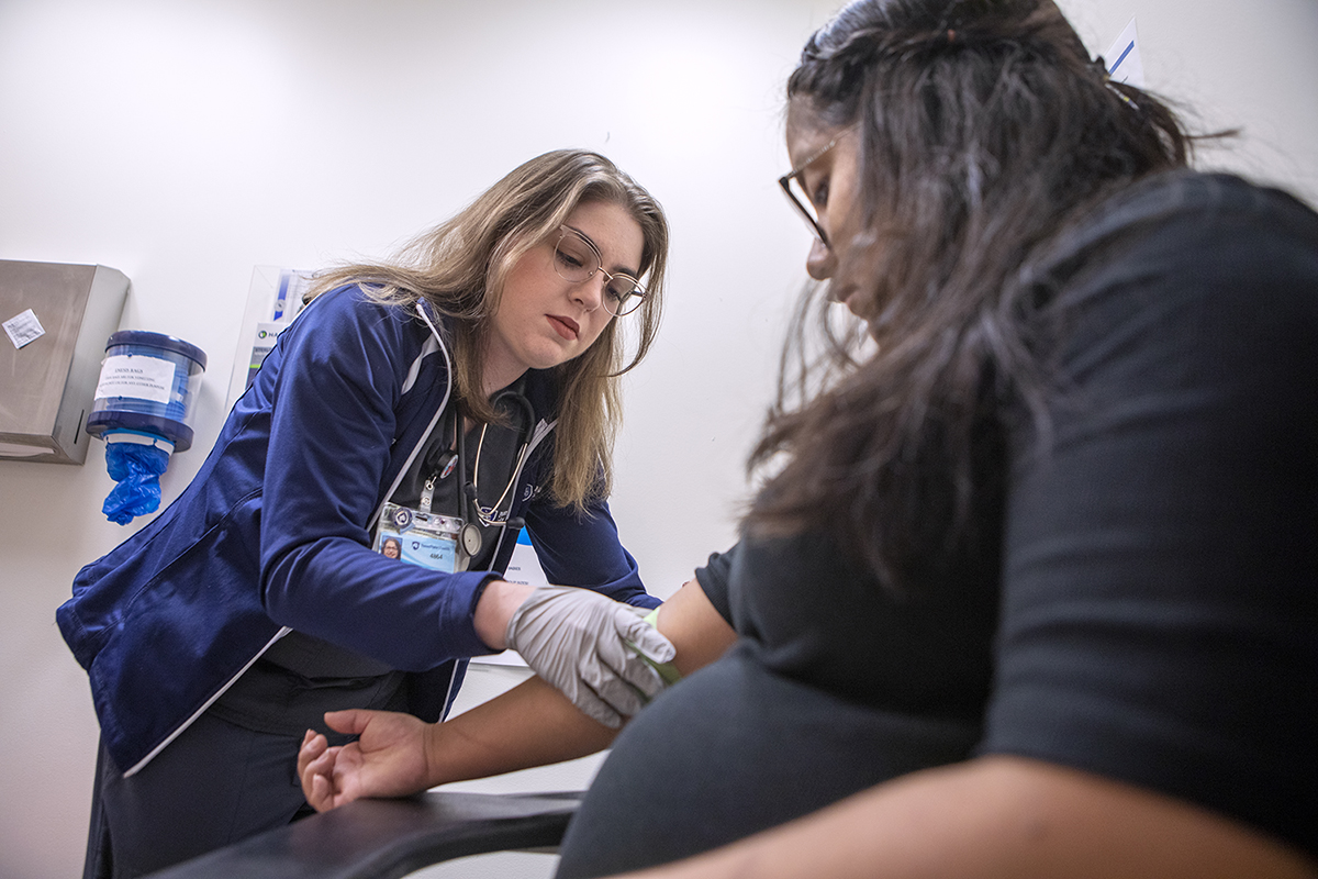 A medical assistant wearing gloves and medical attire works on the outstretched right arm of a patient.