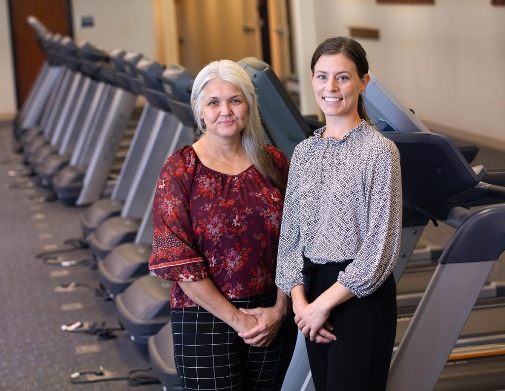 Two women stand in front of a row of treadmills. 