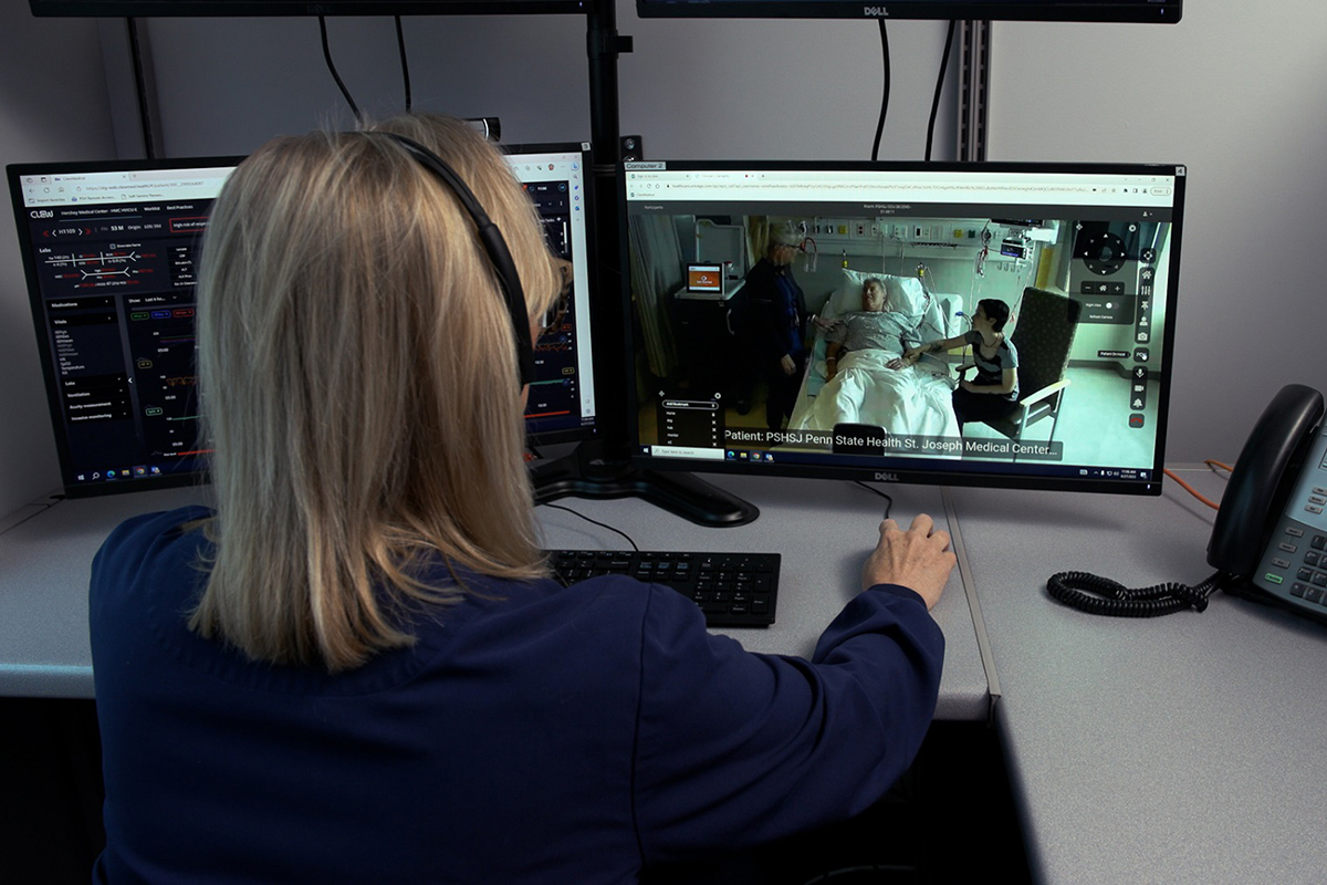 Woman, seen from behind wearing a headset, looks at a video conference on a computer monitor showing a male patient lying in a bed in a hospital room, with a woman sitting beside the bed reaching out to him.