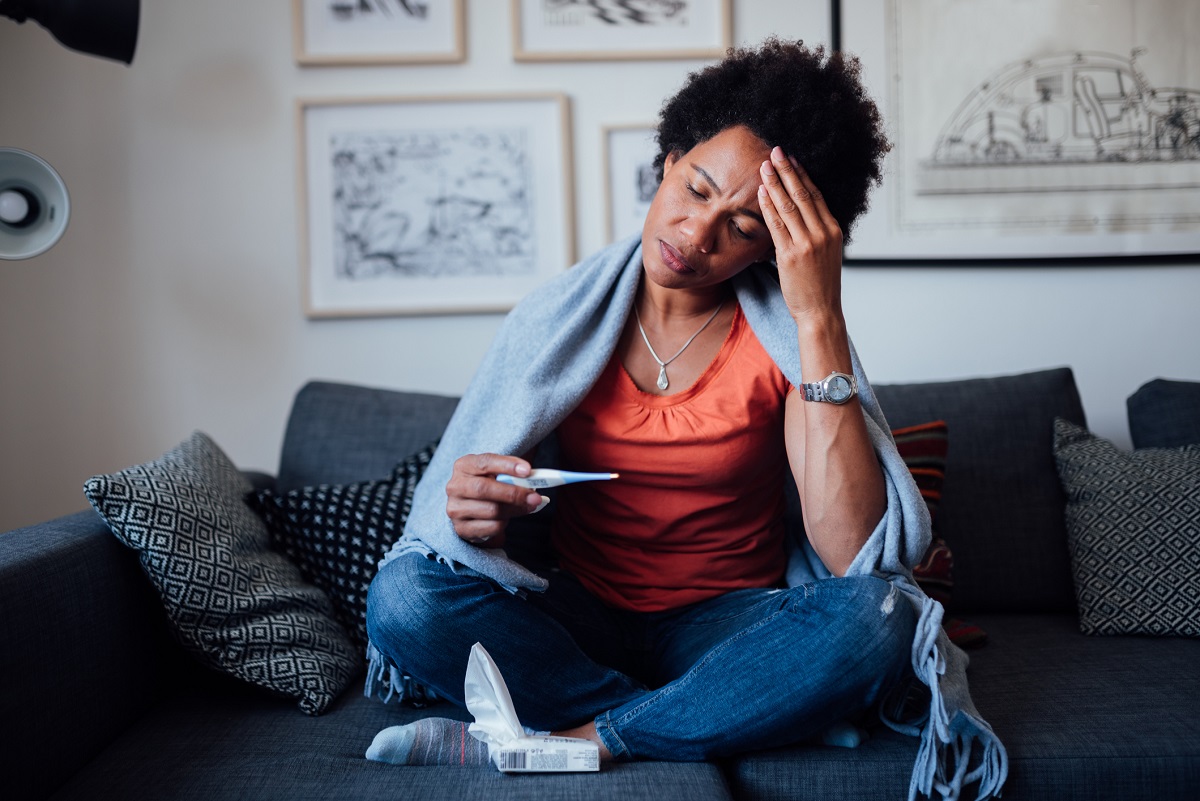 A young woman seated on a couch clutches her forehead and reads a thermometer. A box of tissues is positioned near her lap and a blanket is draped over her shoulders.