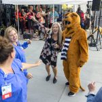 Three people dance with the costumed Nittany Lion. Musicians perform under a tent in the background.