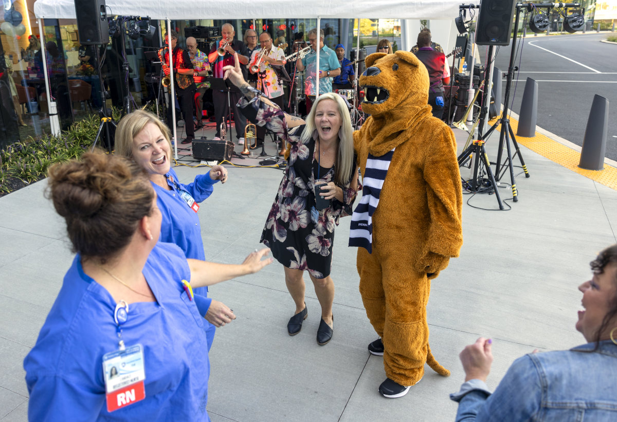 Three people dance with the costumed Nittany Lion. Musicians perform under a tent in the background.