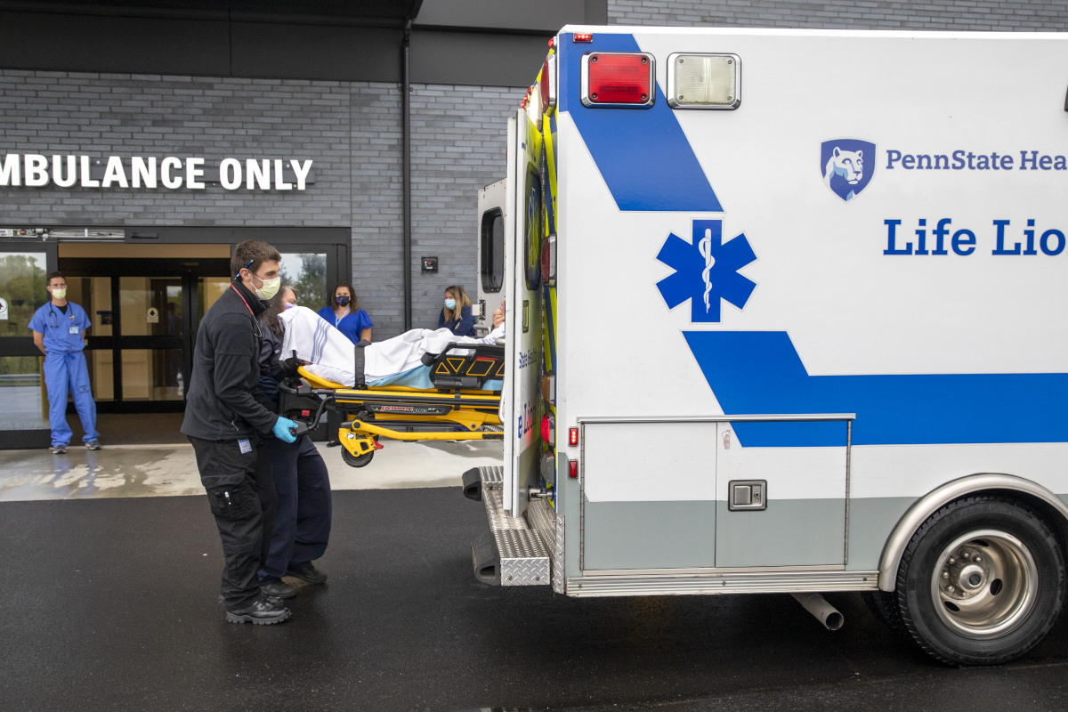 Two individuals remove a litter carrying a patient from the back of an ambulance while three people look on in the background. The words 