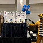 The Nittany Lion mascot gestures toward two displays of the Penn State Health Advanced Practice Providers of the Year awards. He is wearing a striped scarf. The poster boards are on a table, which is on a stage. A podium is to the left, and balloons are in the background.