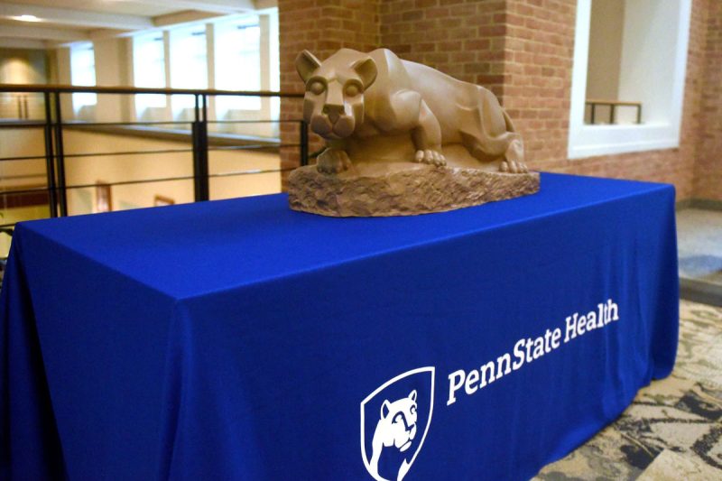A table has a blue table cloth over it that says "Penn State Health." On top of the table is a small Nittany Lion statue.