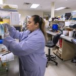 Osiris Martinez-Urquilla holds up a vial in front of lab testing equipment at Penn State Health St. Joseph Medical Center’s laboratory. She is wearing a lab coat and rubber gloves. Her hair is in a ponytail. Behind her is a work bench with laptops and two chairs.