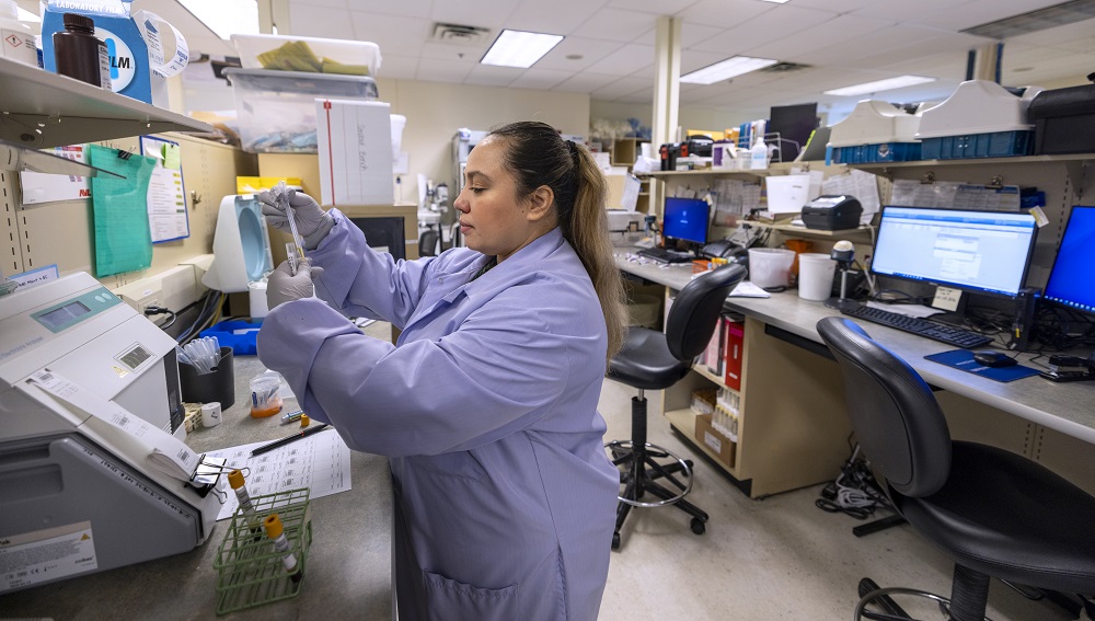 Osiris Martinez-Urquilla holds up a vial in front of lab testing equipment at Penn State Health St. Joseph Medical Center’s laboratory. She is wearing a lab coat and rubber gloves. Her hair is in a ponytail. Behind her is a work bench with laptops and two chairs.