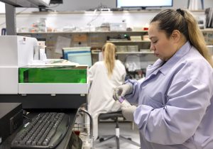 Osiris Martinez-Urquilla looks at a vial she is holding in the St. Joseph Medical Center laboratory. She is wearing a lab coat and rubber gloves. In front of her is a container for medical tests filled with a solution and a computer keyboard. Behind her a woman lab technician is seated on a stool working at a computer.