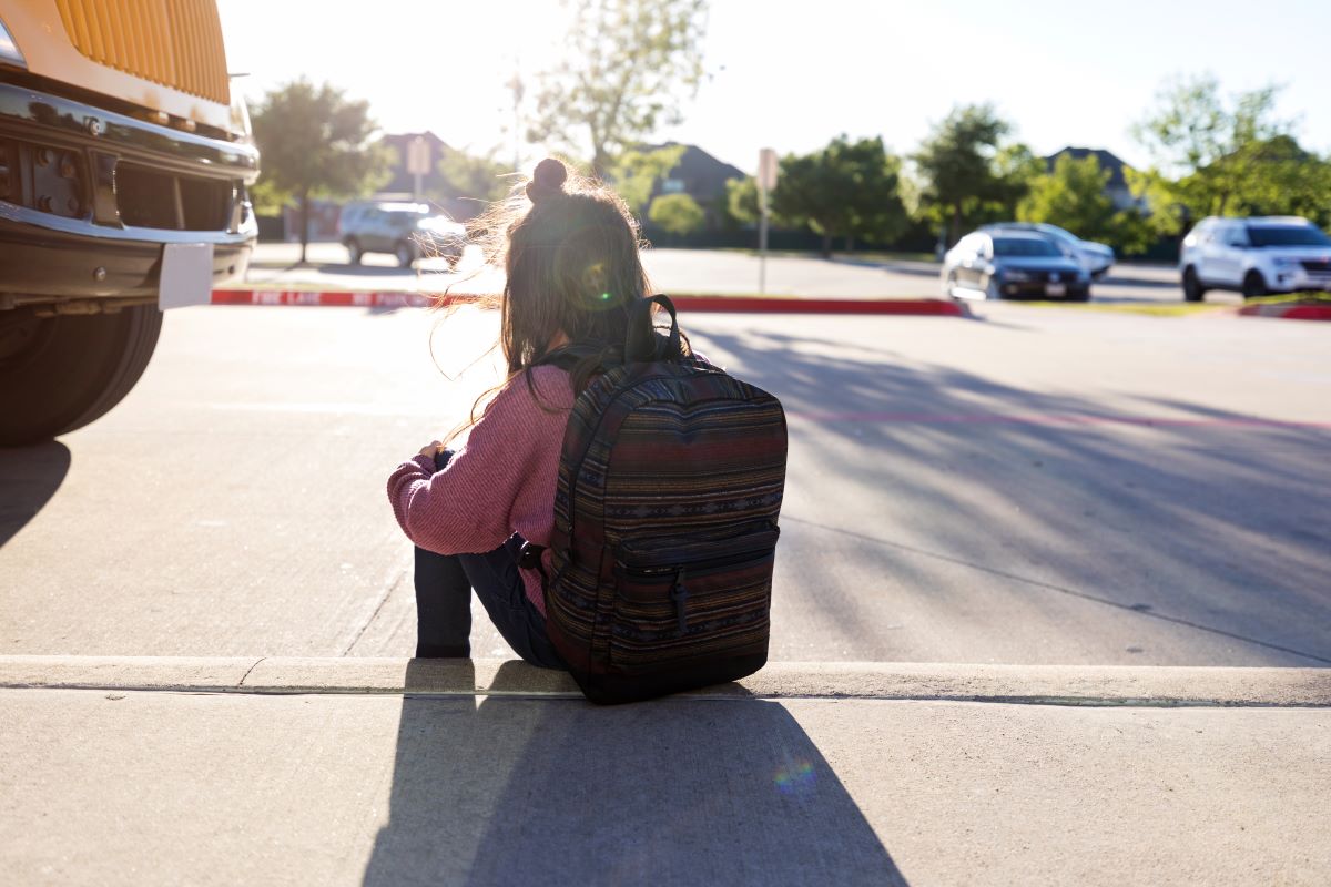 Child sitting on the curb of a road, wearing a backpack, holding knees, looking down, facing away. Front of a school bus on the left. Parking lot in the distant background.