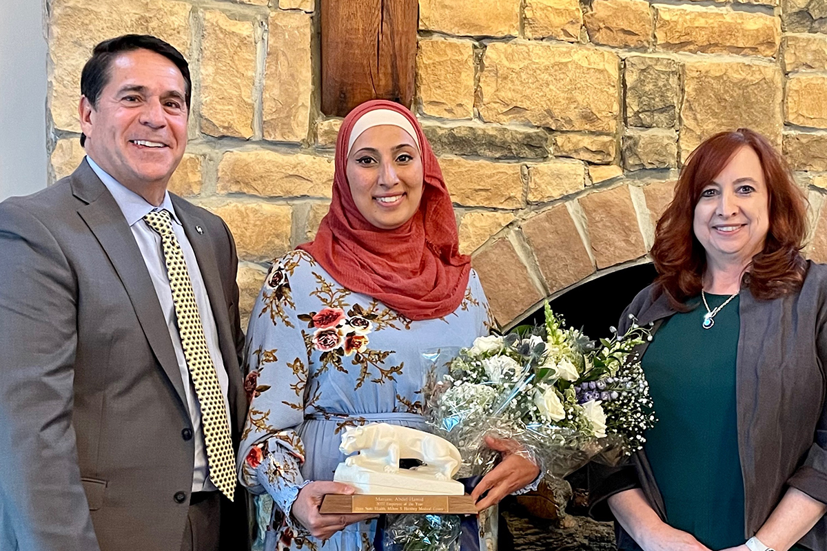 Three people posing in front of a stone fireplace – gentleman on left in suit, smiling; lady in middle, holding a bouquet of flowers and a Nittany Lion statue trophy; lady on right, smiling.